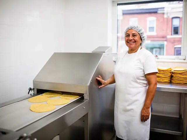 Smiling woman in a hairnet and white apron with her hand on a stainless tortilla conveyor oven